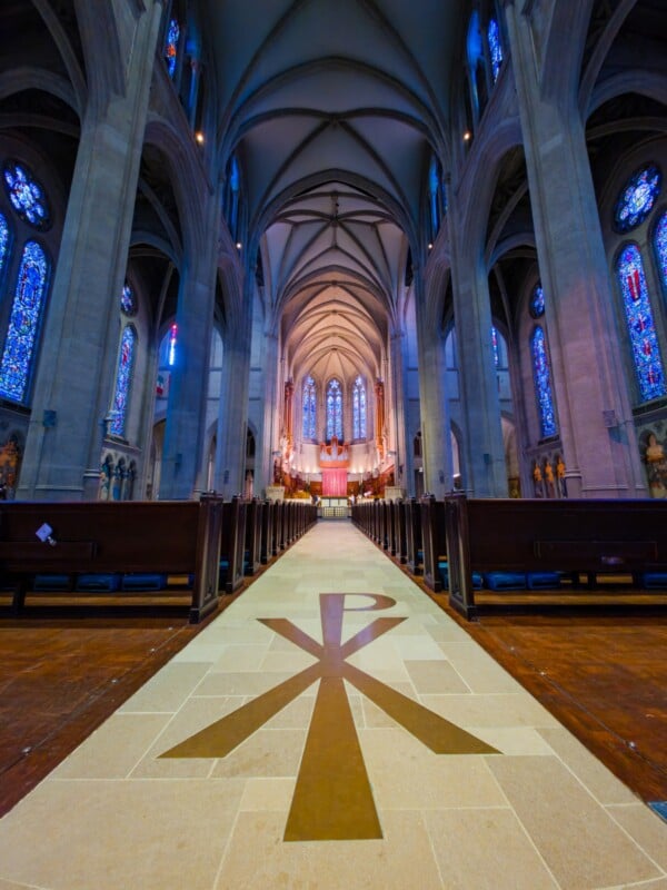 A wide-angle view of a grand cathedral interior with tall arches, stained glass windows, wooden pews, and a Chi Rho Christian symbol on the central aisle leading to an illuminated altar.