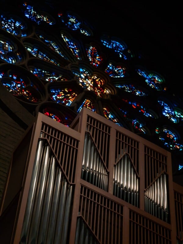A large church pipe organ stands in front of a colorful stained glass rose window, with sunlight shining through the glass and illuminating the dark interior.