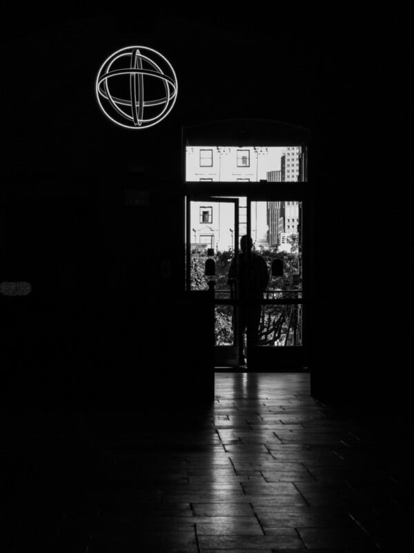 A silhouetted figure stands outside glass doors, with city buildings visible beyond. Inside, a glowing circular symbol hangs above the entrance in the dim, shadowy interior.