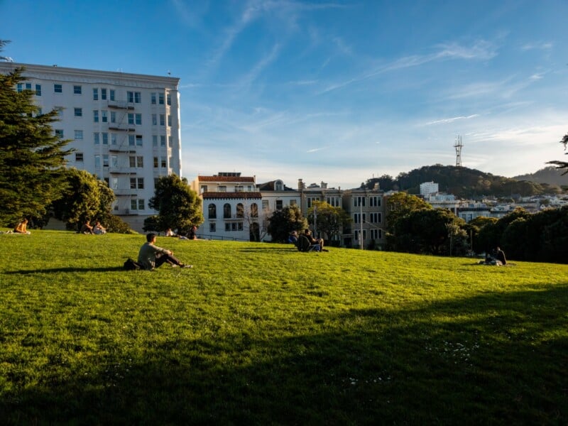 People sit and relax on a grassy hill in a city park during late afternoon. Apartment buildings and trees surround the park, with a communication tower visible on a distant hill under a clear blue sky.