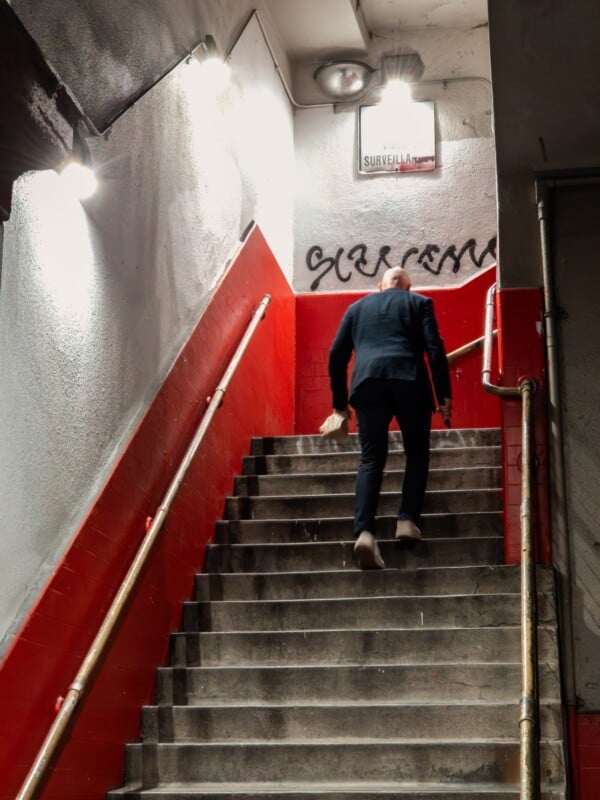 A man in a suit walks up a concrete stairwell with red and white walls, holding something in his hand. The stairwell is lit by overhead lights, and a surveillance sign is visible at the top.