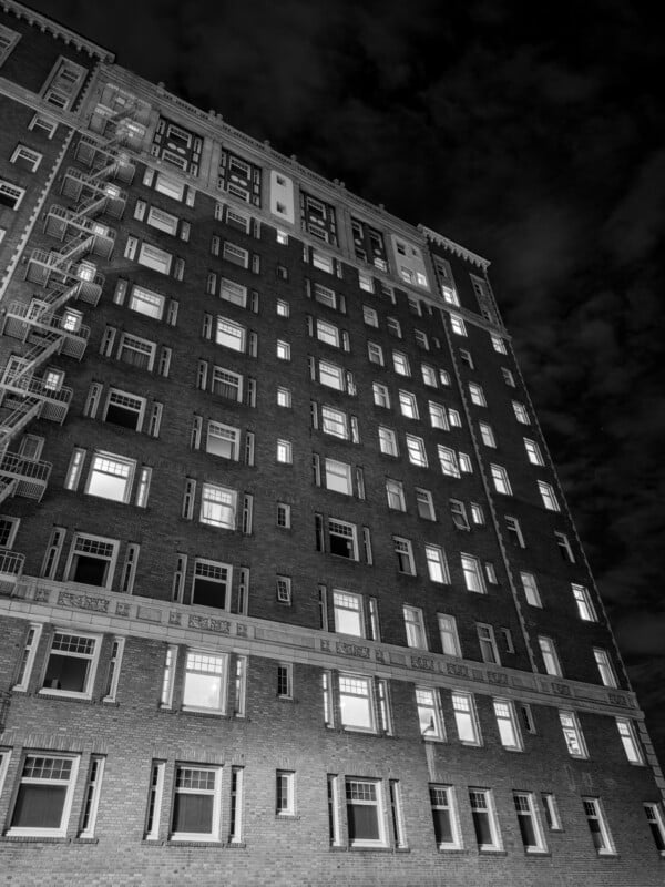 Black and white photo of a tall brick apartment building at night, with many windows lit from inside; a fire escape runs along the left side, and dramatic clouds fill the sky above.