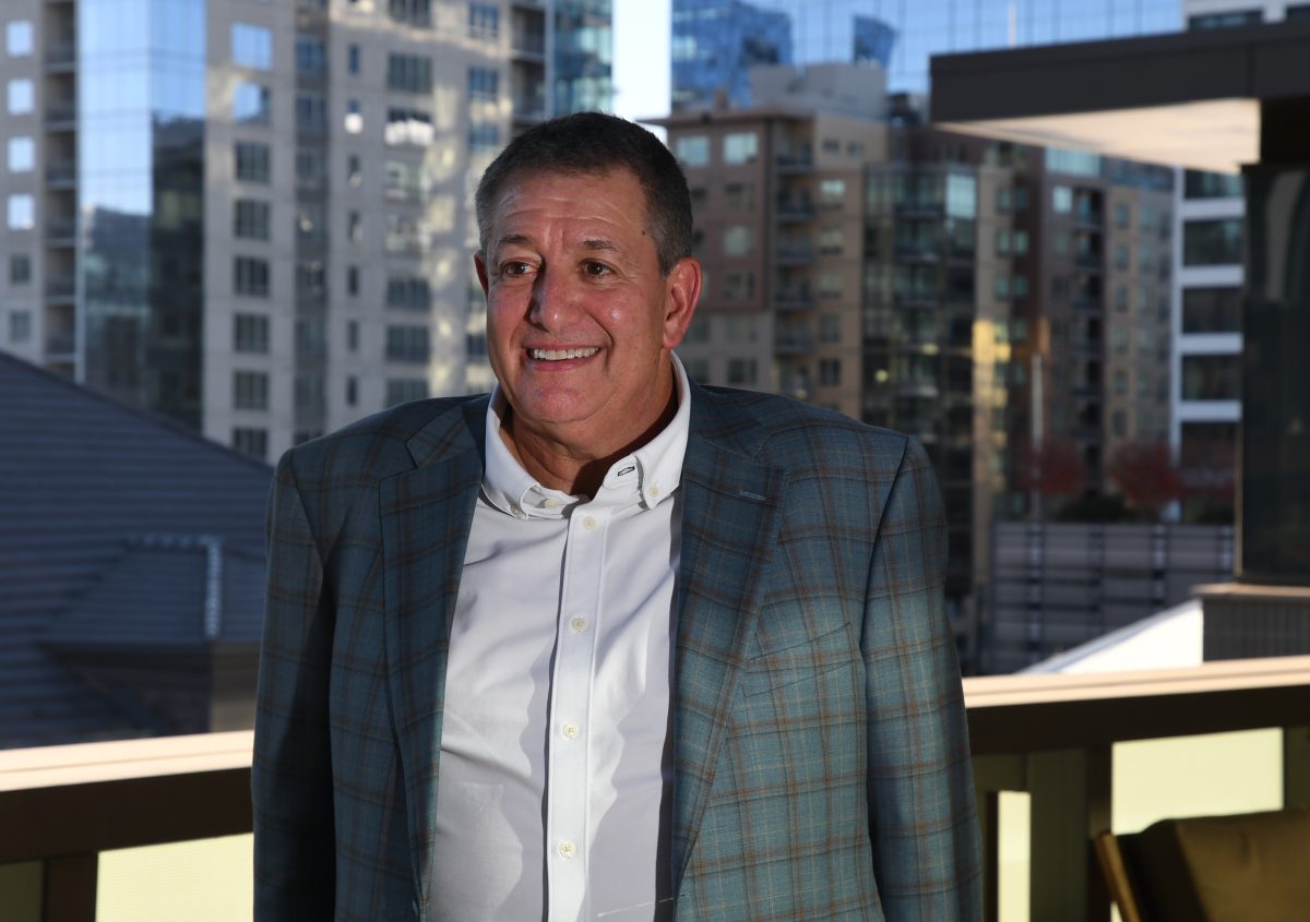 A man in a checked blazer and white shirt stands on a balcony with modern city buildings in the background.
