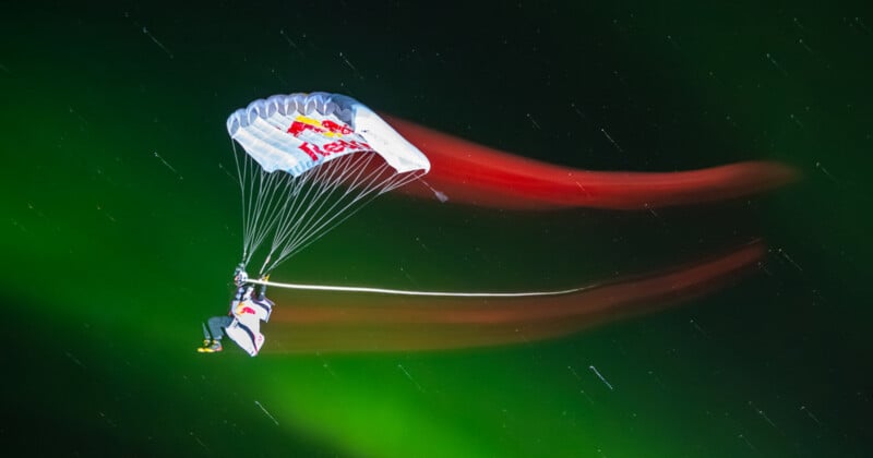 A skydiver with a Red Bull parachute glides through a night sky illuminated by green auroras, with red light trails following behind.