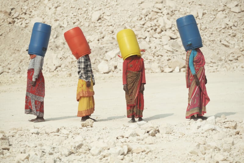 Four women in colorful traditional clothes walk in a line across a rocky, sandy landscape, each carrying a large plastic container on their head. The containers are blue, red, and yellow.