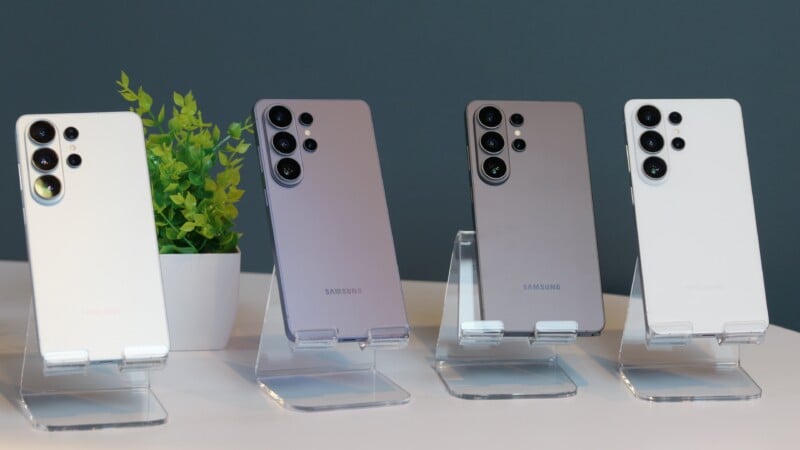 Four Samsung smartphones in different colors are displayed upright on clear stands, with a small potted plant in the background on a white surface.