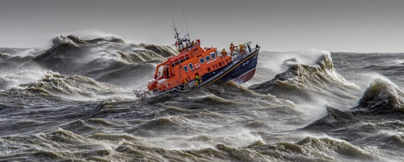 An orange lifeboat battles rough, stormy waves in a dark gray sea, tilted by the force of the water as crew members in yellow gear work on deck.