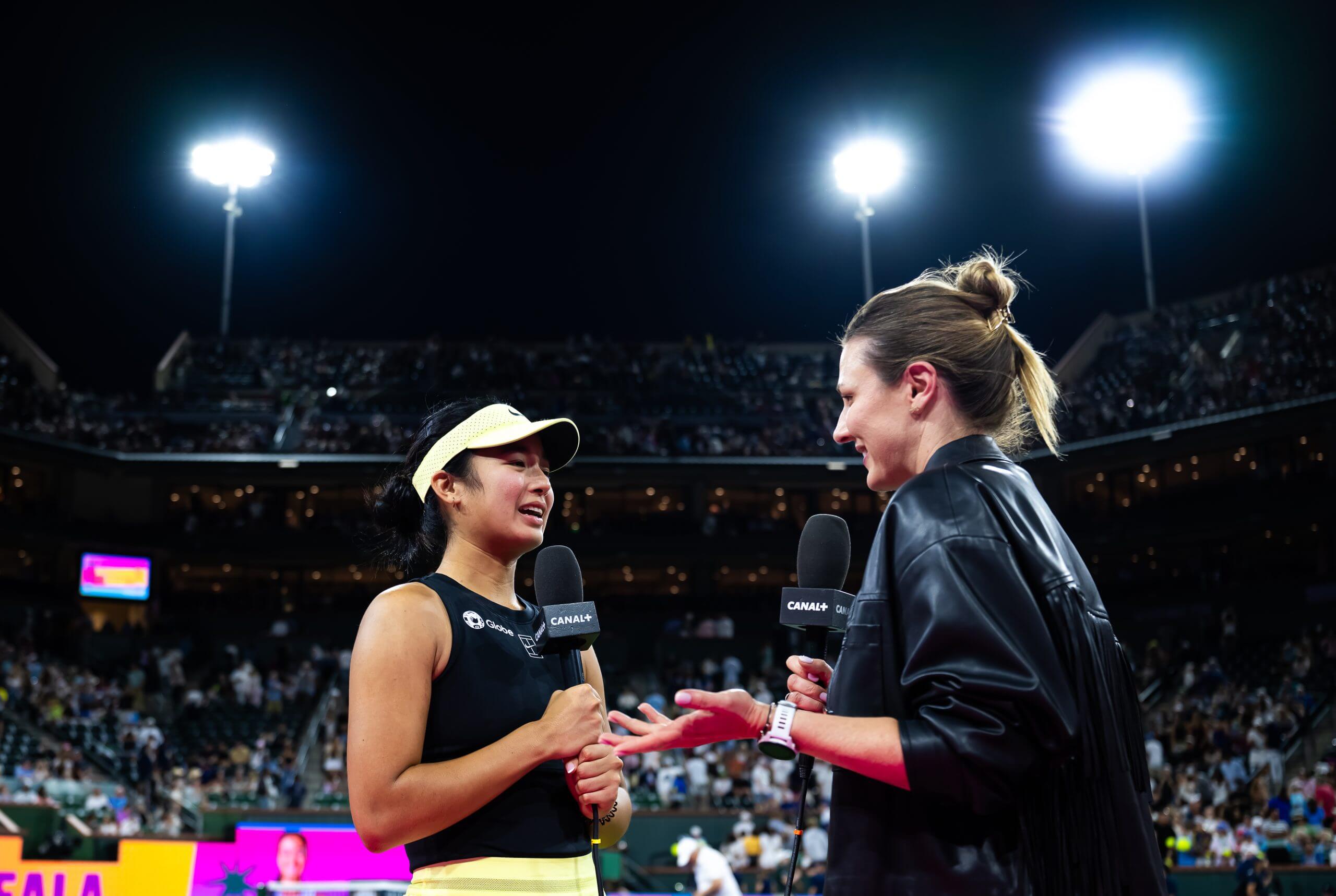 Alex Eala stands holding a microphone in front of a woman wearing a black jacket and also holding a microphone, with crowds and floodlights in the background.