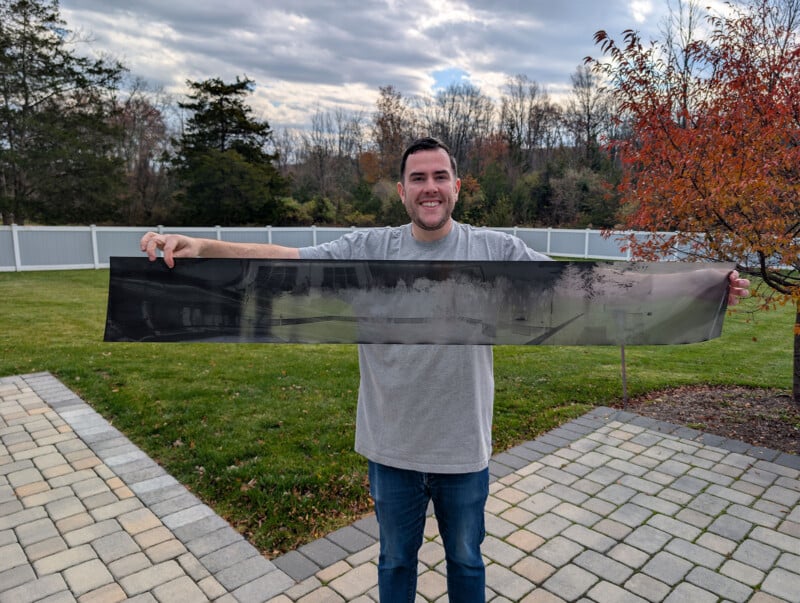 A man stands outdoors on a patio holding a long, rectangular black-and-white panoramic photograph, smiling at the camera. Trees, grass, and a white fence are visible in the background under a cloudy sky.
