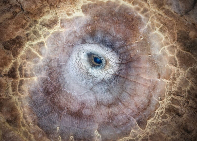 Aerial view of a round, crater-like geological formation with concentric rings, cracks, and earthy tones of brown, tan, and gray, resembling an abstract eye.