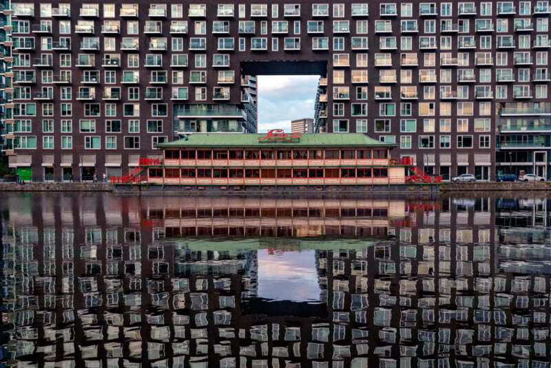 A large modern building with many windows stands by the water, featuring a large rectangular opening in the center. A red and green houseboat is docked in front, both reflecting clearly in the calm water below.