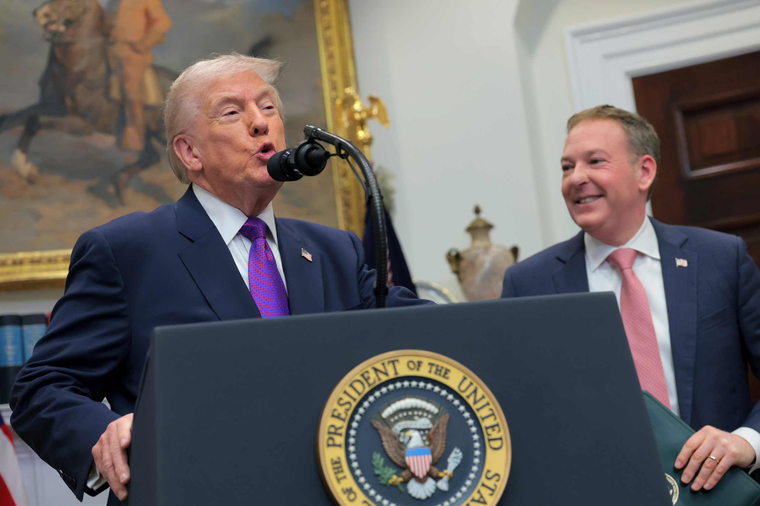 Donald Trump, an elderly man wearing a suit and purple tie, speaks at a lectern. Next to him a middle aged man wearing a red tie smiles.