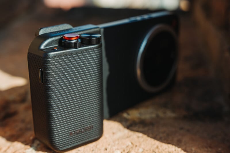 A close-up of a modern digital camera with a textured grip and a prominent red shutter button, resting on a rough stone surface in sunlight.
