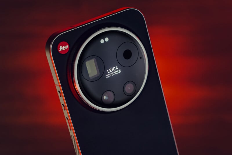 A close-up of a smartphone with a large circular Leica camera module and lens, set against a warm red blurred background. The phone has a sleek black and metallic design.