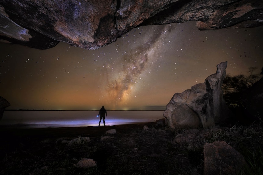 The Milky Way rises over a lake, with a man standing in silhouette at the lake's edge, as seen from a cave in the foreground.