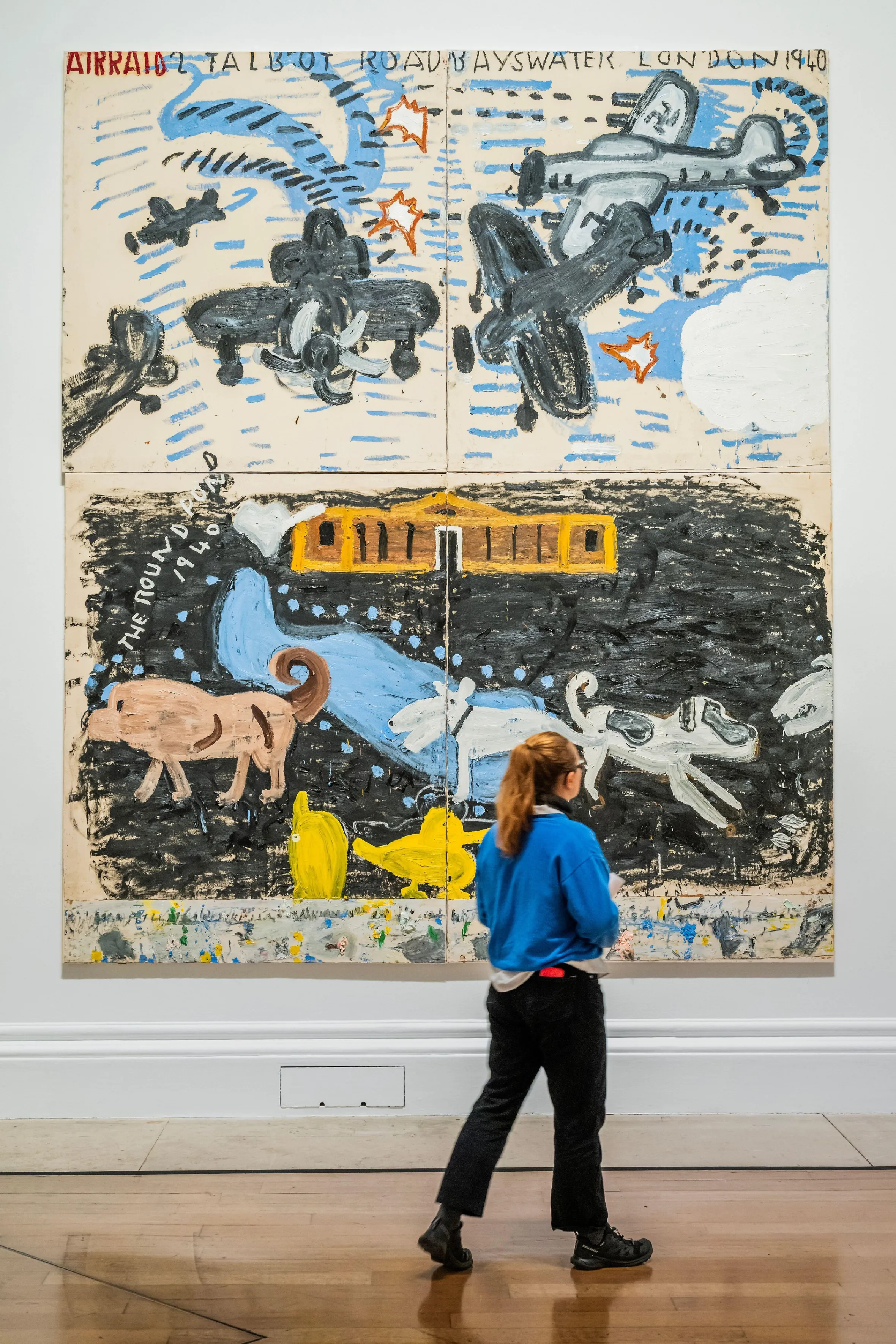 Illustration by Rose Wylie titled "Park Dogs & Air Raid, 2017" showing planes, dogs, and a building, with a person viewing the artwork.