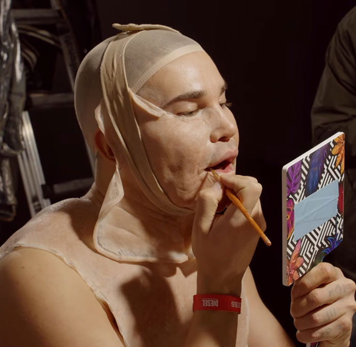Drag queen applying makeup while wearing a mask to pose as Jim Carrey in a behind-the-scenes setting. Drag queen applying makeup while wearing a mask to pose as Jim Carrey in a behind-the-scenes setting.