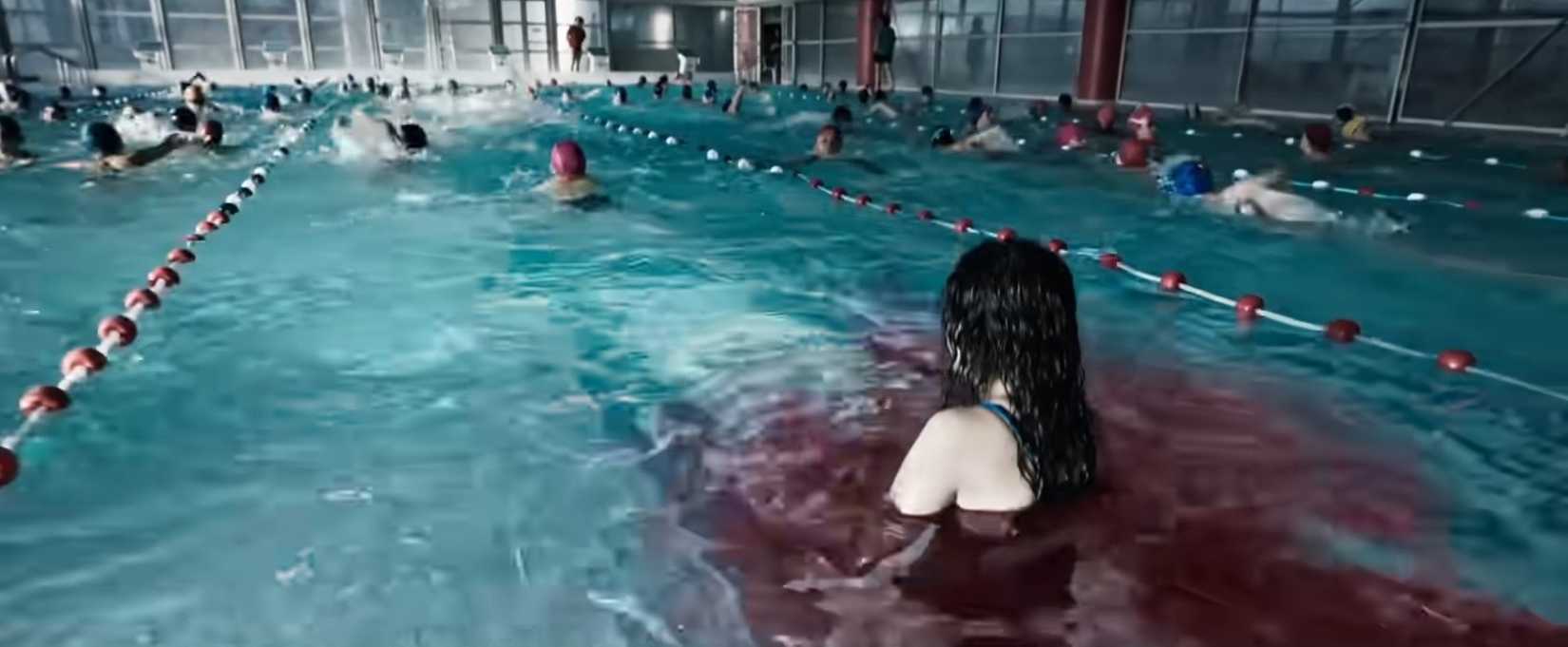 A girl with her back to the camera stands in a school swimming pool, the water around her becoming cloudy with blood, with the camera positioned slightly above her, in a scene from Alpha (2025).