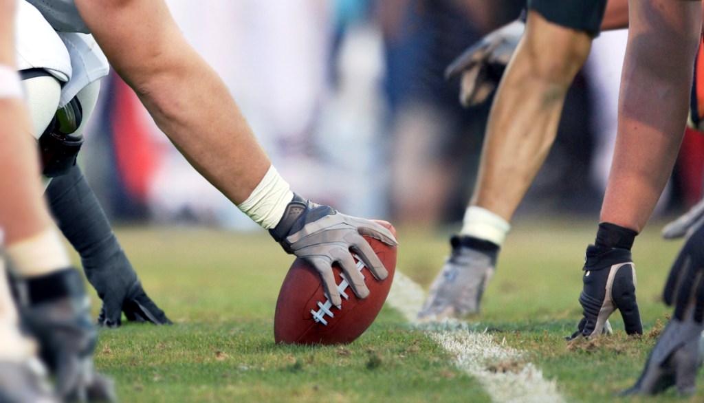 An American football player’s gloved hand on the ball at the line of scrimmage, with other players in position on the grass.