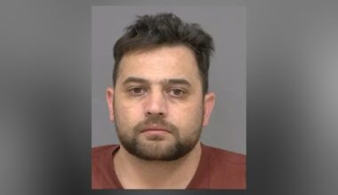 Close-up of a man with short dark hair and a trimmed beard, looking straight at the camera with a neutral expression against a plain background.