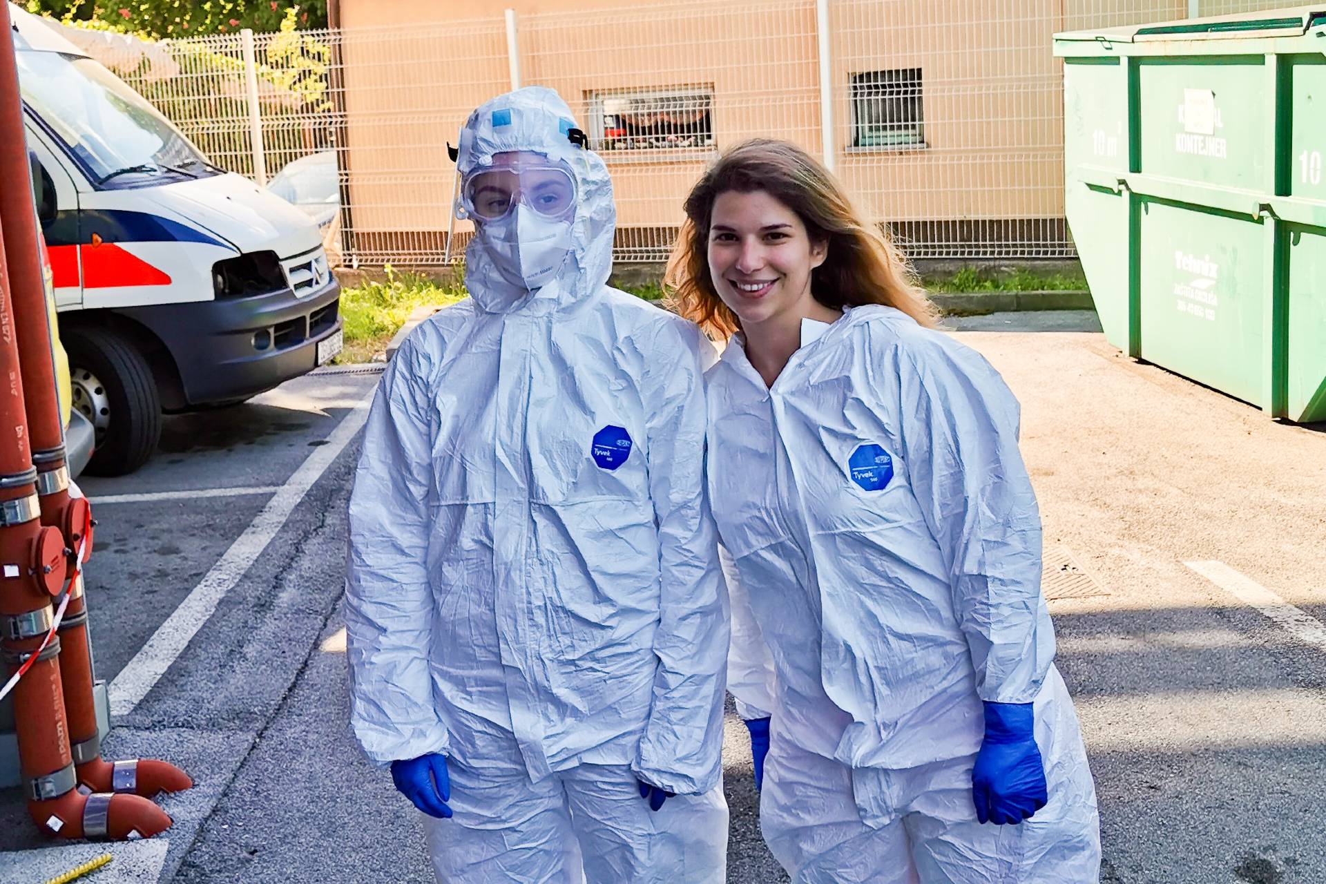 Two healthcare workers in protective suits standing outside near an ambulance.
