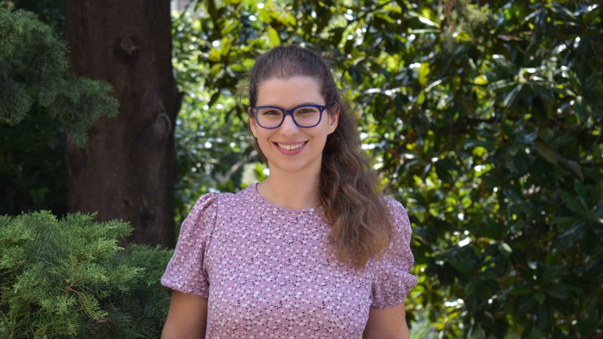 A smiling woman with glasses standing outdoors in front of green trees.