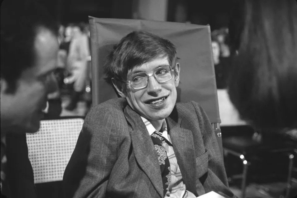 Black and white photo of Stephen Hawking smiling at someone to his side