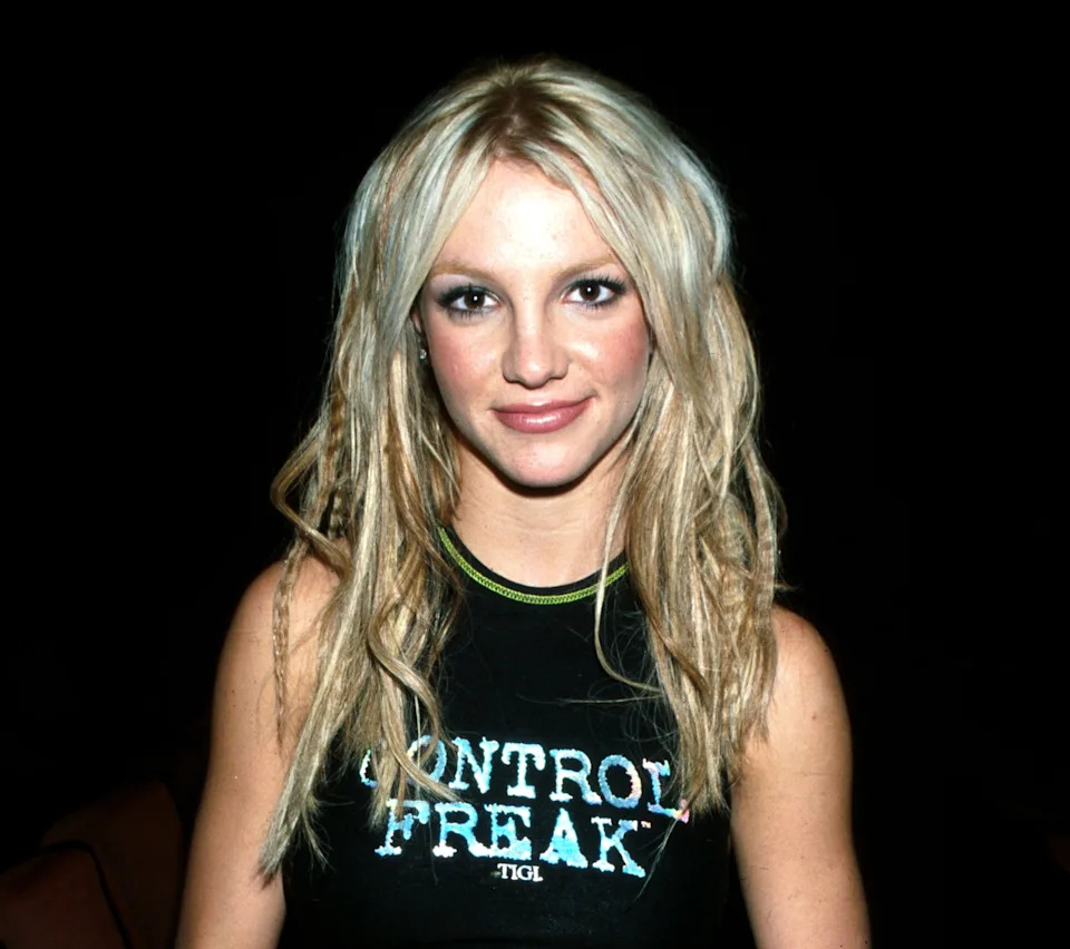 Britney Spears wearing a "Control Freak" crop top and hip-hugging pants, with long hair and a navel piercing, standing indoors