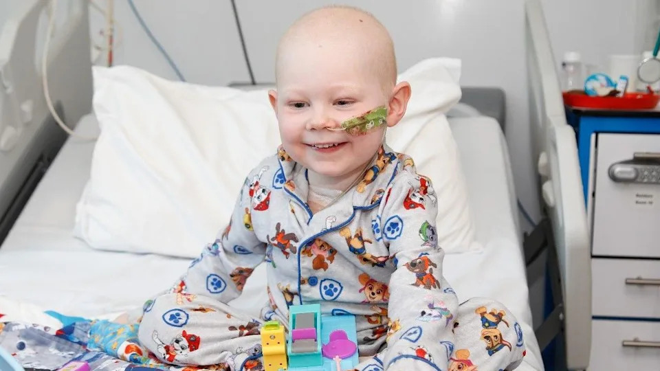 Vinnie, 4, is sitting on a hospital bed. 
He has no hair and has a tube going into his nose. 
Vinnie is smiling and wearing grey and blue pajamas with dogs on them. They are the dogs from a children's TV programme called Paw Patrol.
He has a few toys in front of him on the bed.