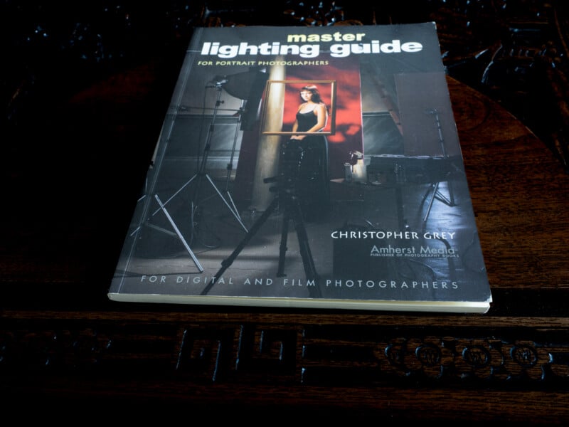 A book titled "Master Lighting Guide for Portrait Photographers" by Christopher Grey rests on a dark, ornate wooden surface. The cover shows a photography studio setup with lights and a portrait subject.