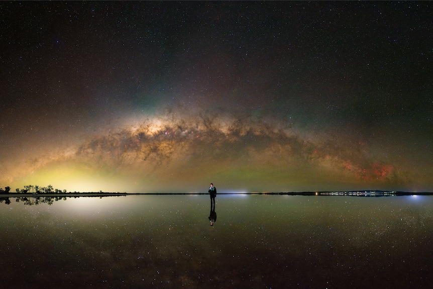 A man stands on a dry lake in the middle of the image with the entire milky way above him.