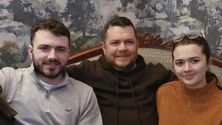 Mark is pictured with his son and daughter. To his left is is his son Danny, who has bearded and has short, wavy hair. Mark's left is his daughter, whose fair is tied back from here  face. She wears an orange jumper and she has reading glasses on her head. 