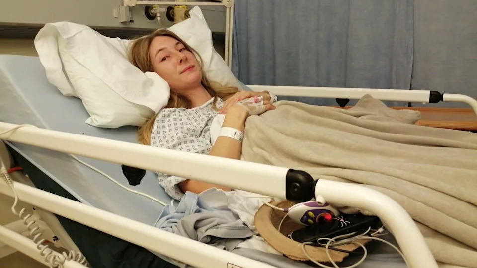 The image shows Iona Hall lying in a hospital bed wearing a white gown, looking at the camera.