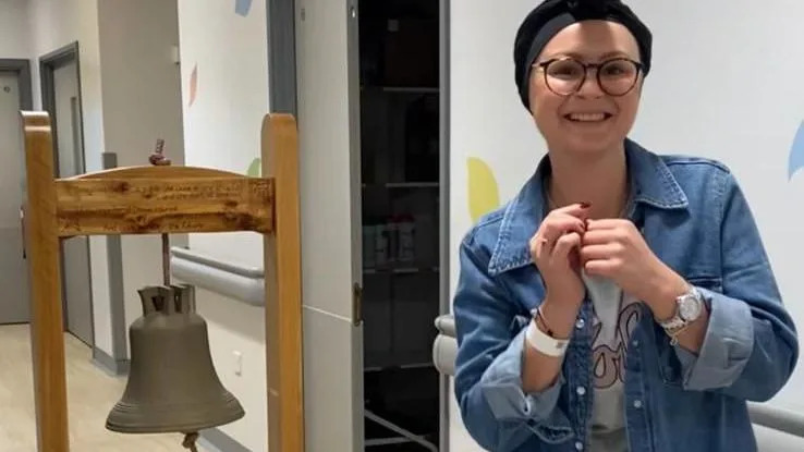Jo Owens stands to the right of the picture, smiling and ringing the celebratory bell to signify being cancer clear. She wears a denim jacket and has wrapped a piece of dark fabric over her head.