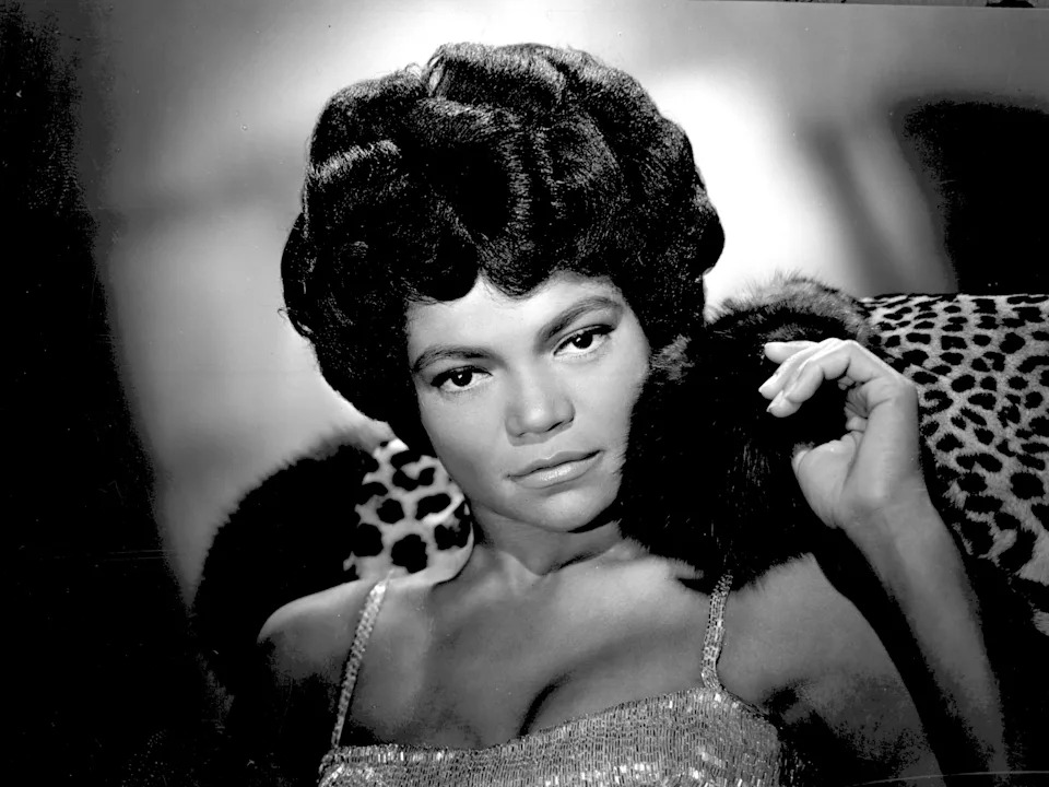 Eartha Kitt in a shimmering dress with an elegant, fur-trimmed coat seated against a leopard-print backdrop