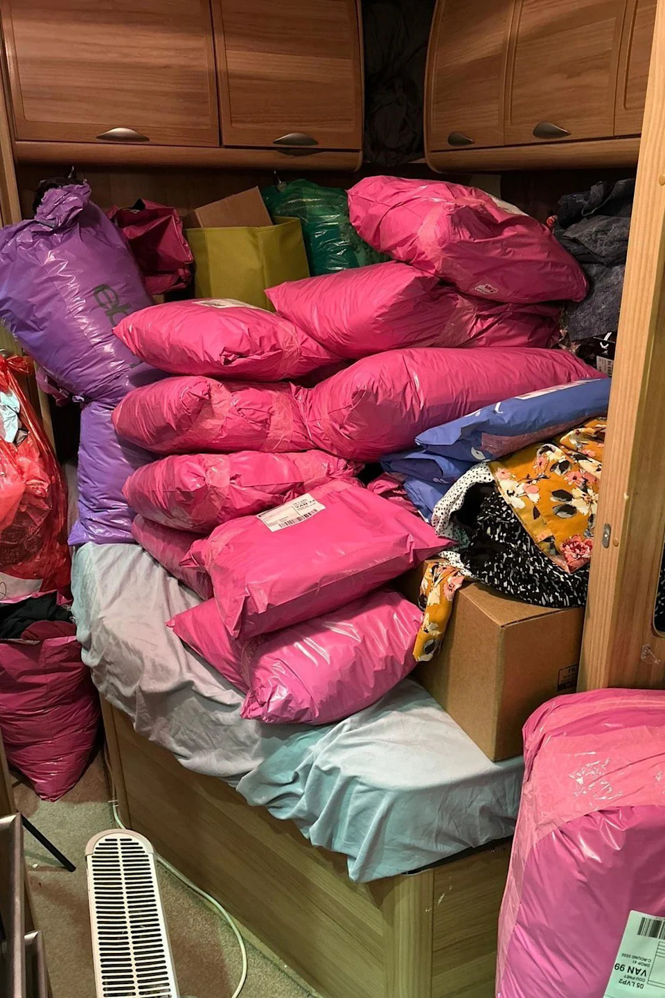 Stacks of pink parcels can be seen piled on top of a double bed within a caravan. The parcels pile up to the bottom of overheard cupboards, which are a dark wood colour. 