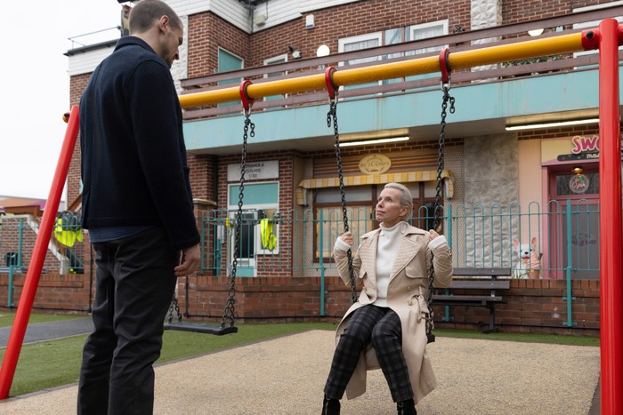 corrie-carl-debbie-swings Debbie sitting on a swing talking to a standing Carl in Coronation Street