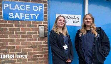 Two women in all-blue uniforms stand outside a blue door which says 'Place of Safety'. A brick wall is to the left of the image.
