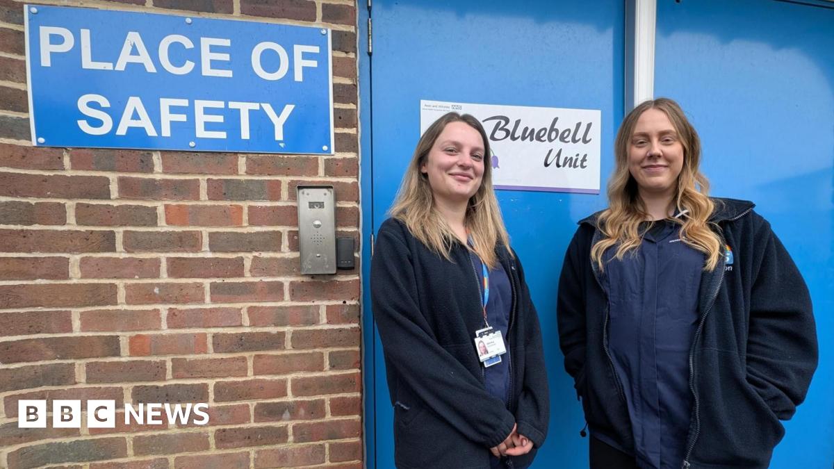 Two women in all-blue uniforms stand outside a blue door which says 'Place of Safety'. A brick wall is to the left of the image.