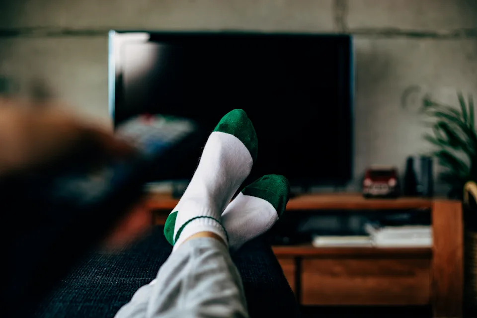 A POV photo of a person laying down on a couch with their legs propped up, turning on a TV with a remote.