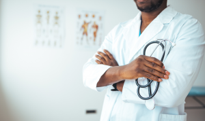 A dark-skinned doctor in a lab coat, holding a stethescope. Their face is cut off above the chin in the photo.