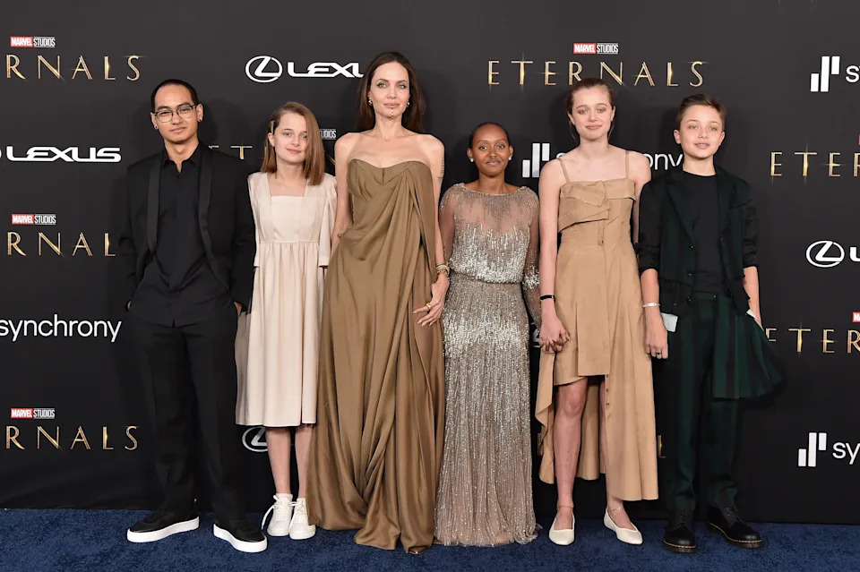 A group on a red carpet in elegant attire attending the premiere of "Eternals," posing for photos. The style is flowing and fashion-forward