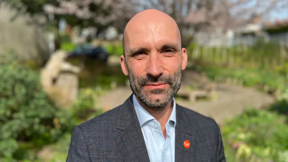 A man perhaps in his 50s stands in a park with a circular patio area behind him. He is bald and has short salt and pepper facial hair. He is wearing a dark checked suit and a light blue top, with a red badge on his lapel.