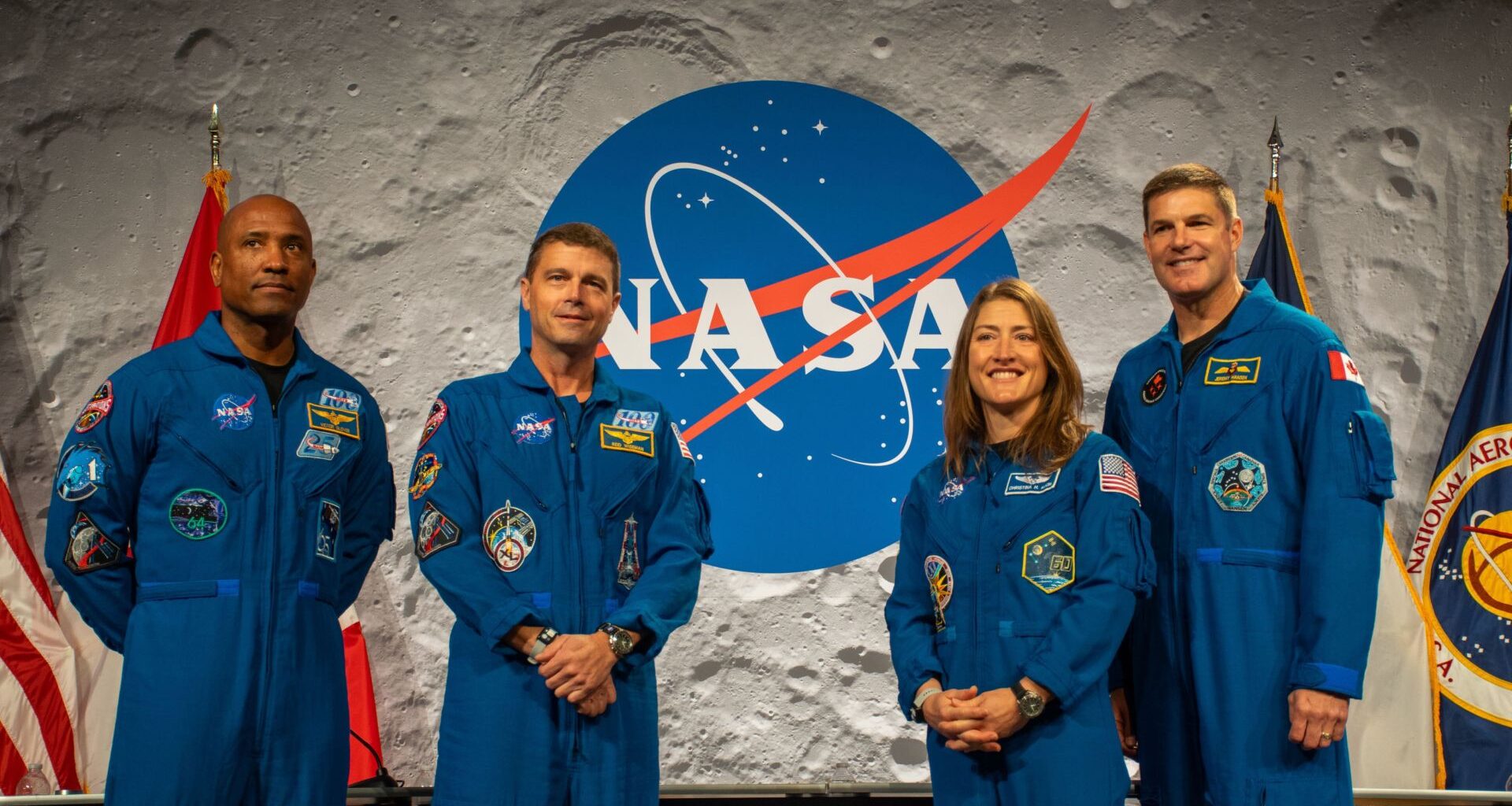 Four people wearing blue jumpsuits stand next to each other in front of a desk with a NASA logo behind them