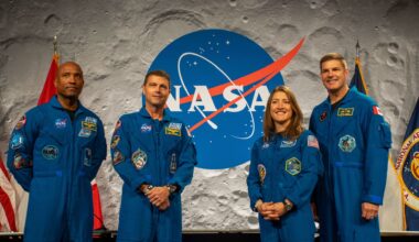 Four people wearing blue jumpsuits stand next to each other in front of a desk with a NASA logo behind them