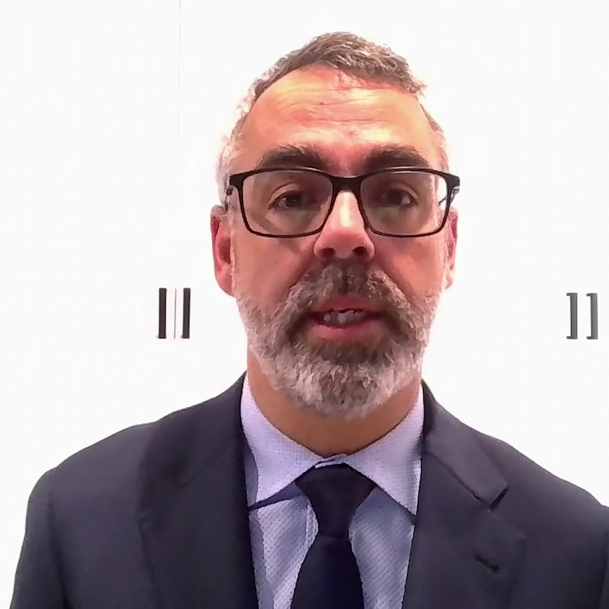 A man in a suit and glasses sits in front of white cupboards