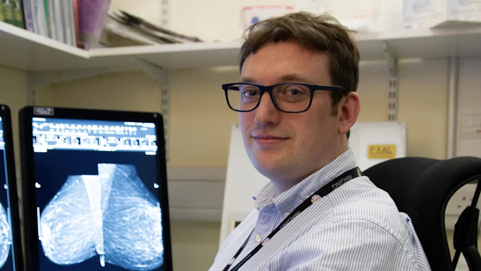 Dr James Tanner is off-centre to the right. He is sitting on a black chair at computers which are showing a breast screening image. He has short brown hair and is wearing black framed glasses, a blue striped shirt and a black lanyard.