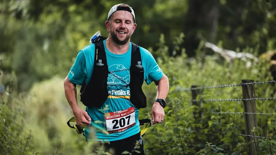 A man in a blue t shirt running with a backpack on and wearing a cap, he has a number on which is 207.