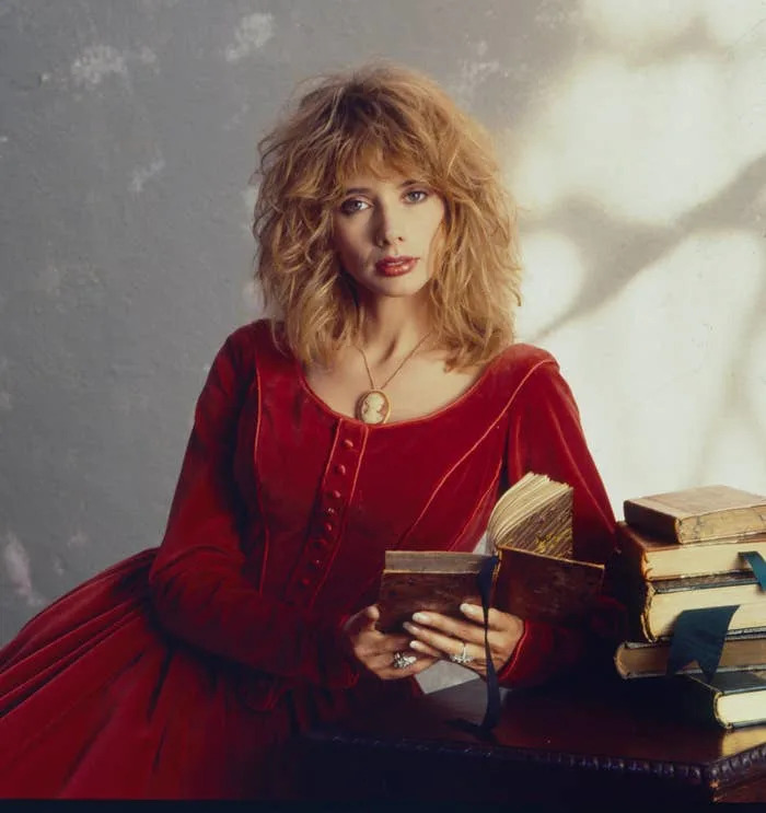 Rosanna Arquette in vintage-style dress reads a book, with a stack of books beside her