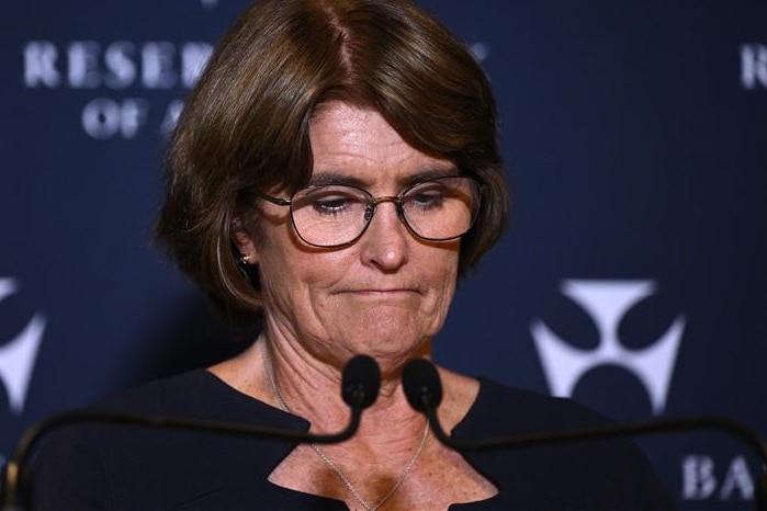 A dark-haired woman in glasses casts her gaze downward as she stands at a lectern in forrnt of an RBA backdrop.